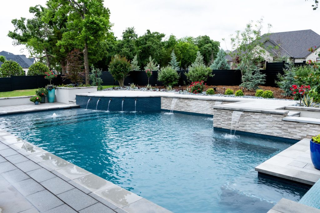 Geometric swimming pool with raised stone wall and sheer descent water features in Oak Tree East, Edmond Oklahoma, featuring Midnight Blue ecoFINISH interior, charcoal Lueder limestone coping, and Platinum Gray paver deck.