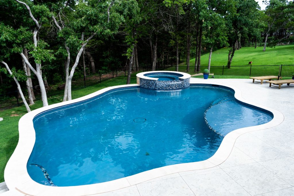 Freeform swimming pool with raised spa in The Falls, Jones Oklahoma featuring Mediterranean Blue ecoFINISH interior and Adobe travertine coping.