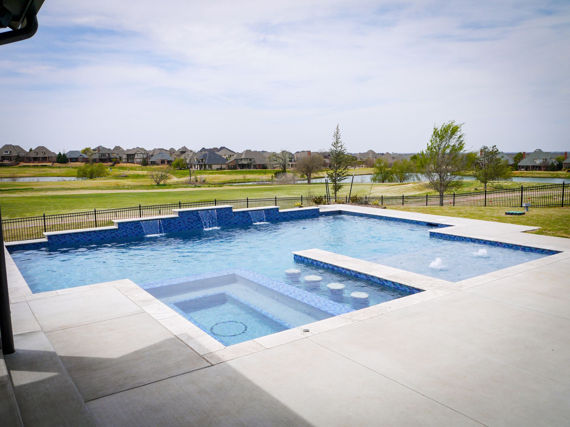 Flush spa connected to geometric swimming pool with wide deck and open backyard view. In-water barstools and sheer descent fountains.