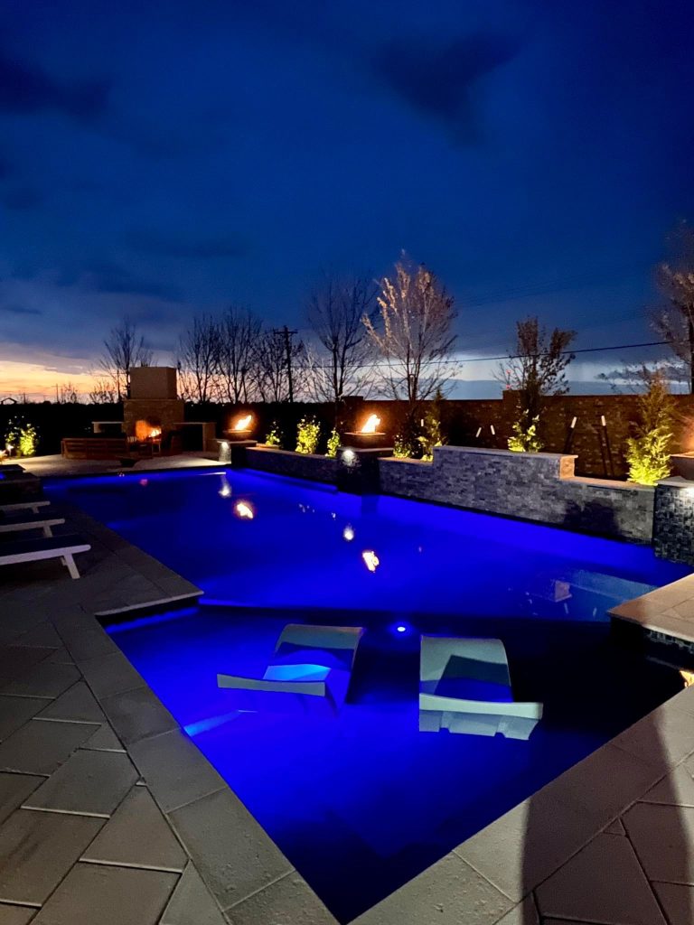 Night view of geometric pool with raised spa and fire bowls in Oak Tree East Edmond Oklahoma featuring Mediterranean Blue ecoFINISH interior and limestone paver deck.